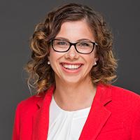 Studio mid-shot of the Hon Amanda Rishworth MP, wearing a red jacket with a white top, black frame glasses and smiling happily at the camera.