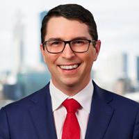 Mid-shot of Patrick Gorman MP, wearing a dark blue suit, white shirt, red tie and black frame glasses, smiling genuinely at the camera.